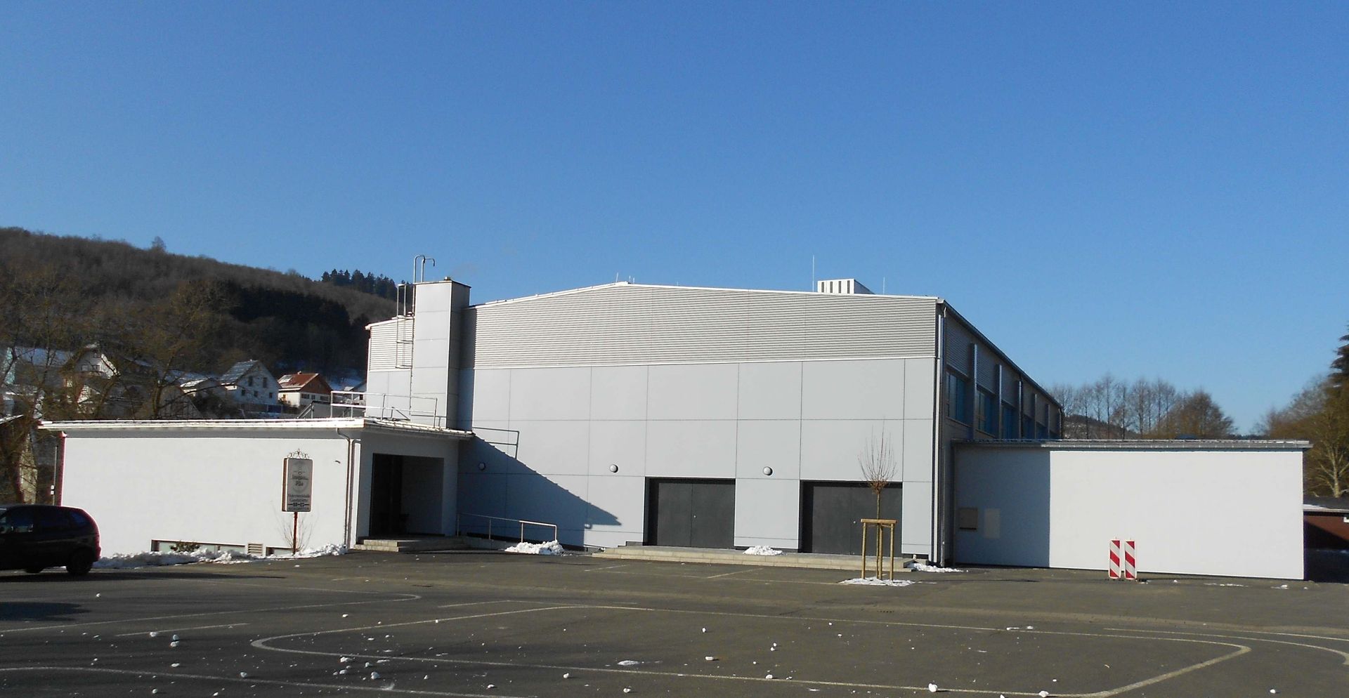 Ein graues, fensterloses Industrie- oder Geschäftsgebäude mit Flachdach unter klarem blauen Himmel, hinter einem Parkplatz.