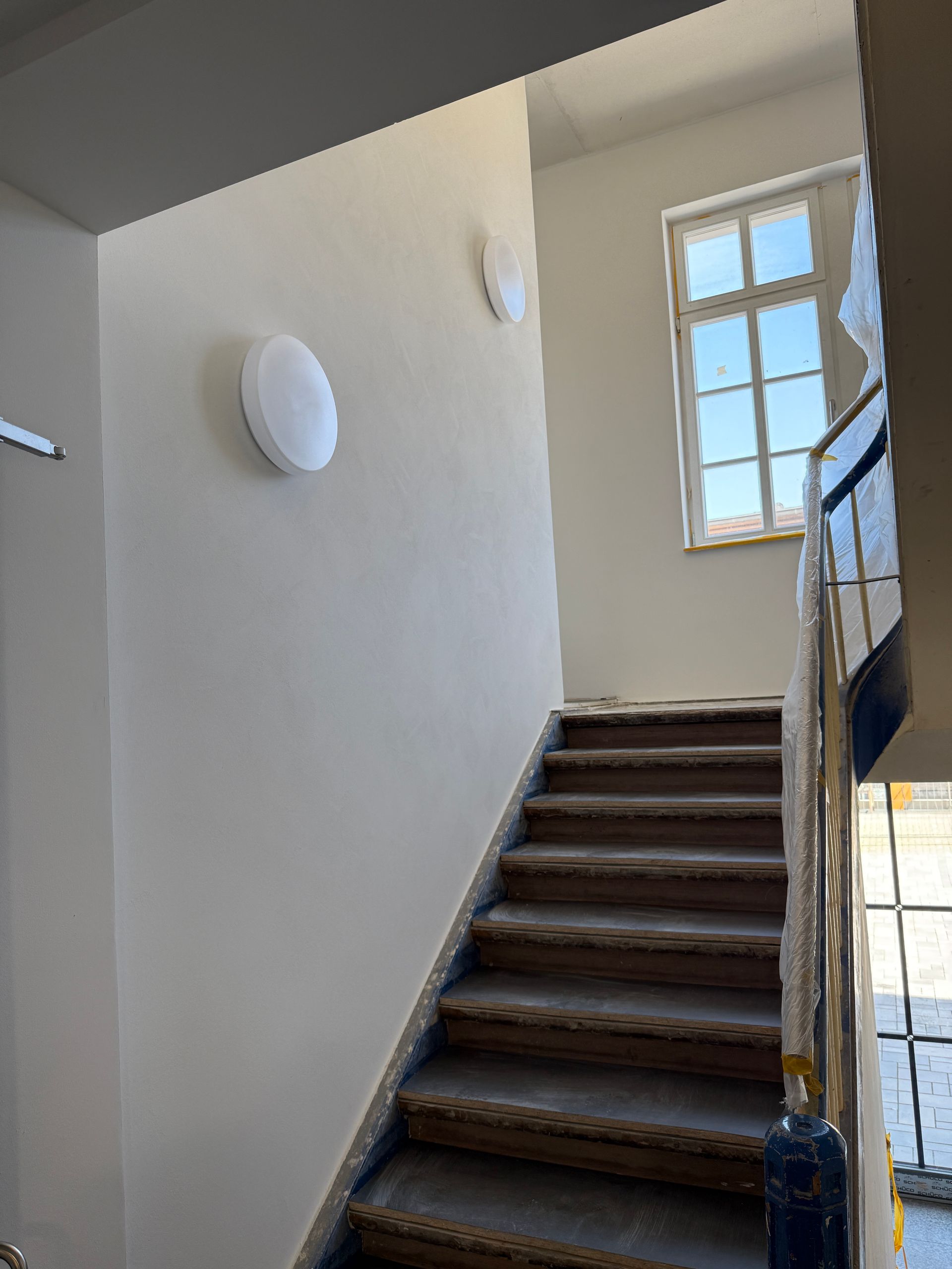 Eine Innentreppe führt zu einem Treppenabsatz mit Fenster, an dessen heller Wand zwei runde Wandleuchten angebracht sind.
