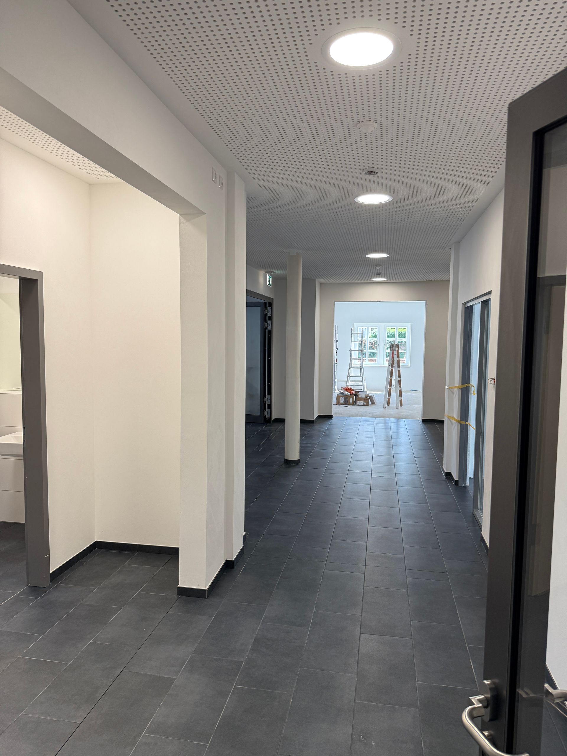 Ein moderner Flur mit dunkel gefliesten Böden, weißen Wänden und einer gemusterten Akustikdecke mit runden Einbaustrahlern.