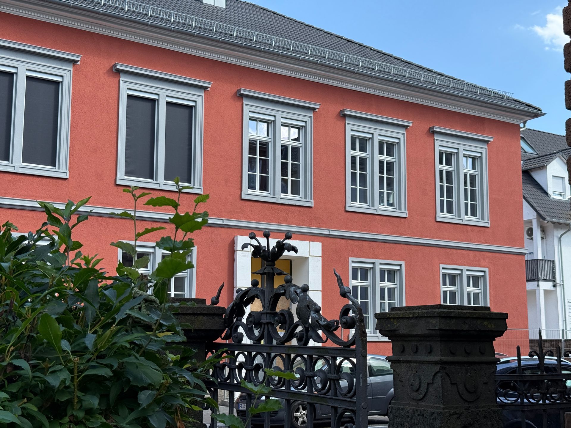 Im Vordergrund ein zweistöckiges, korallenfarbenes Gebäude mit grau umrandeten Fenstern und einem dunklen Eisentor.