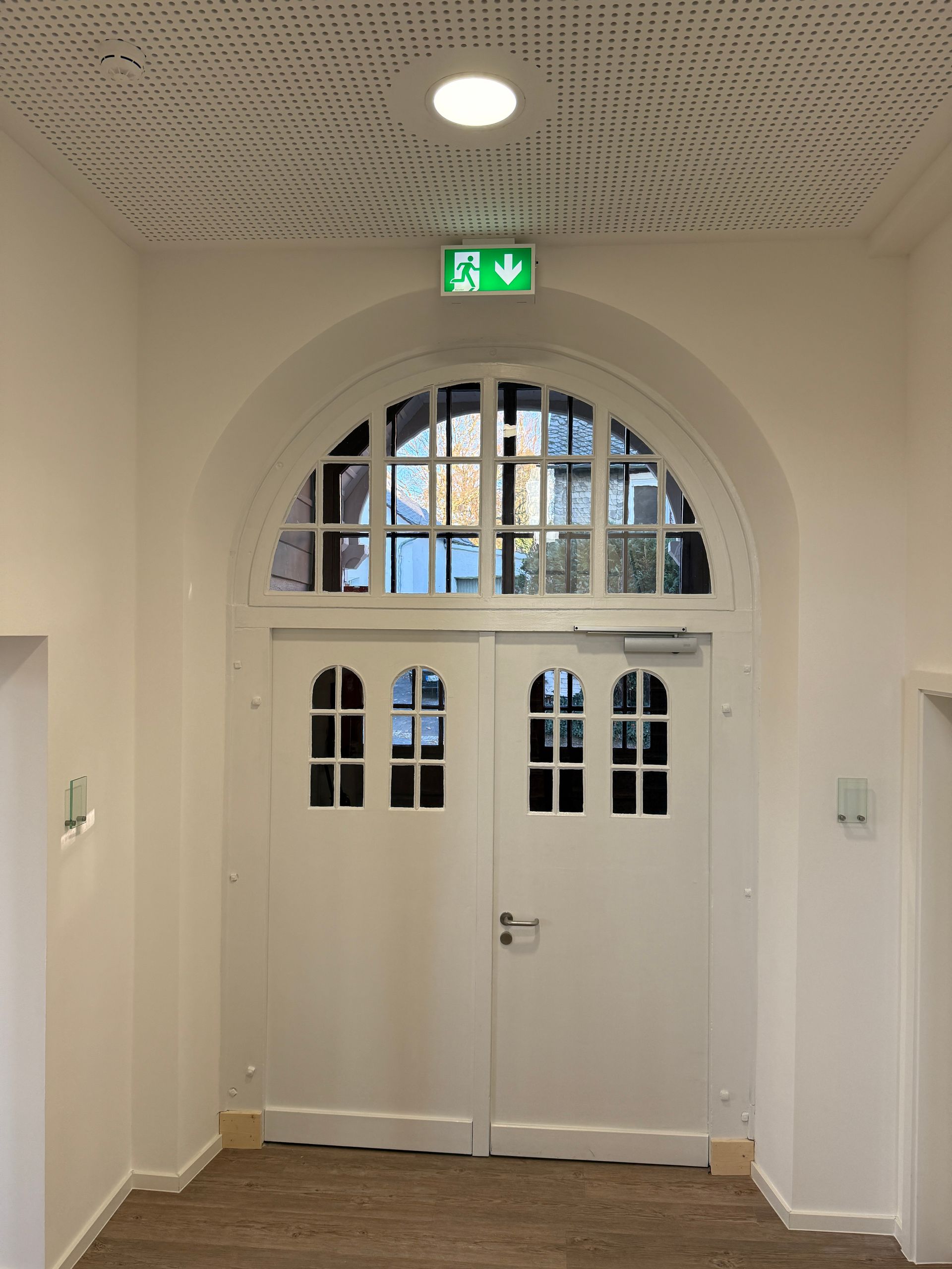 Weiße Doppeltüren mit Glasfenstern unter einem Bogenquerbalken und einem beleuchteten grünen Notausgangsschild.