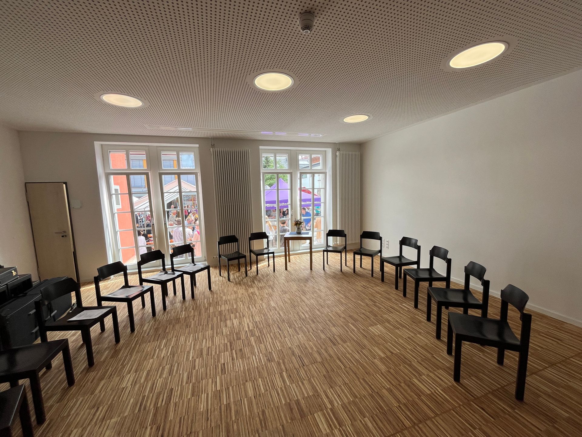 Ein leerer Raum mit einem Kreis schwarzer Stühle auf einem Holzboden, gegenüber zwei Fenstern 