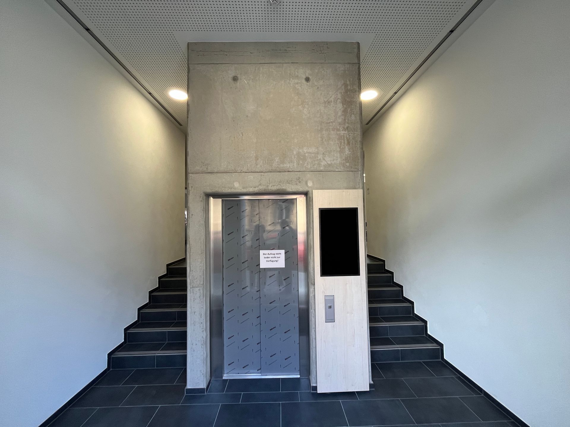 Ein Aufzug in einem Betonschacht zwischen zwei Treppenhäusern, mit einem Schild an der Tür und einem Bedienfeld an der Seite.