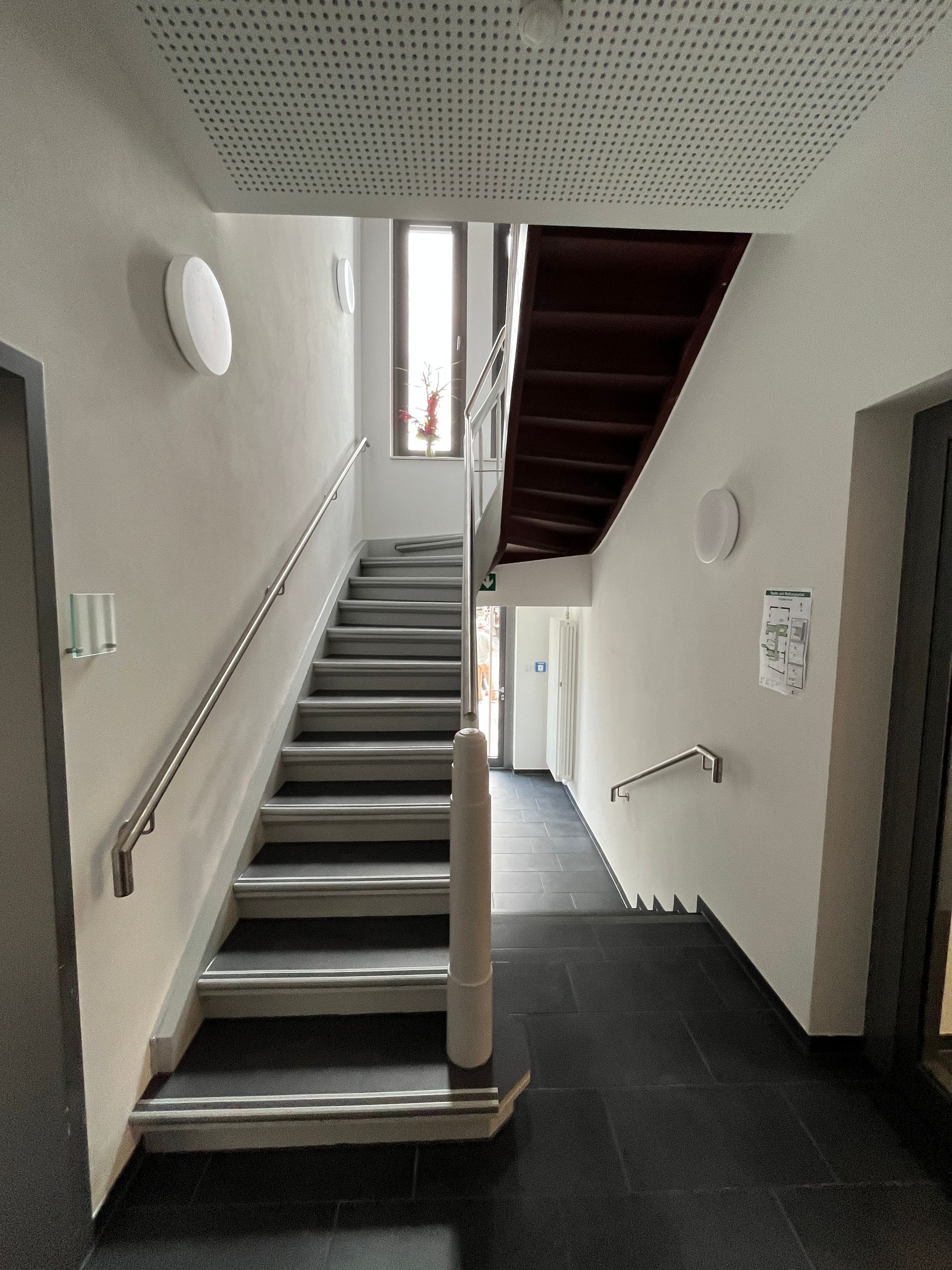 Ein Treppenhaus mit grauen Stufen, silbernem Geländer, hellen Wänden und einem kleinen Fenster im obersten Treppenabsatz.