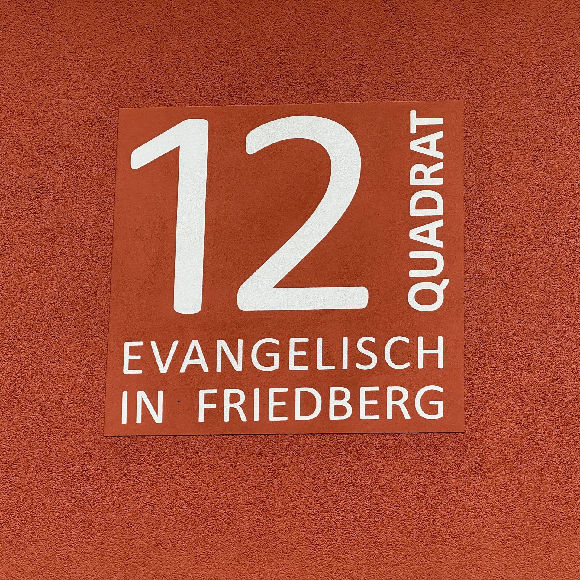 Ein quadratisches, rötlich-braunes Schild mit weißer Aufschrift „12 QUADRAT“ 