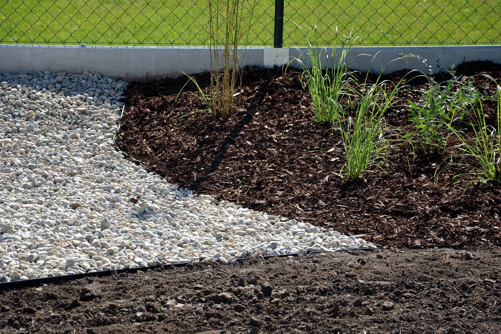 a garden with gravel and mulch and a chain link fence in the background .