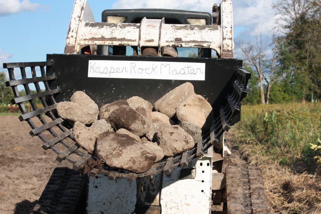 Bobcat skid-steer loader scooping large rocks from dirt; sky and trees in background.