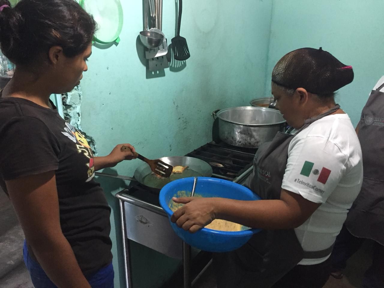 Two women are cooking in a kitchen and one has a mexican flag on her shirt