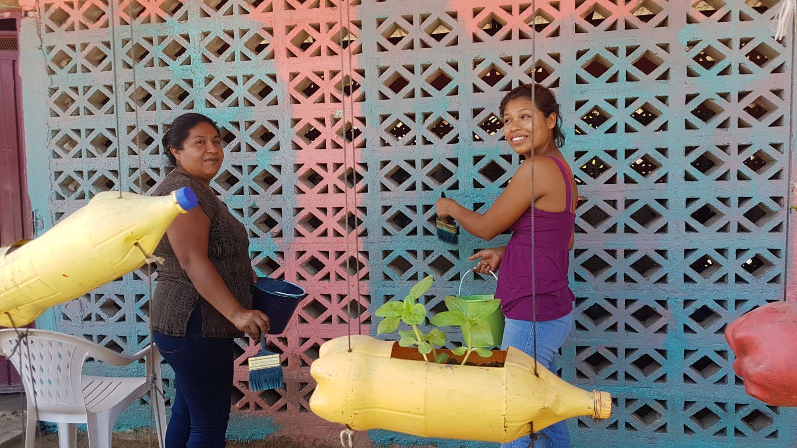 Two women are standing next to each other holding plants in plastic bottles.