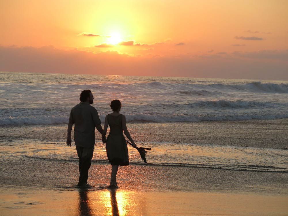 A man and woman are walking on the beach at sunset holding hands.