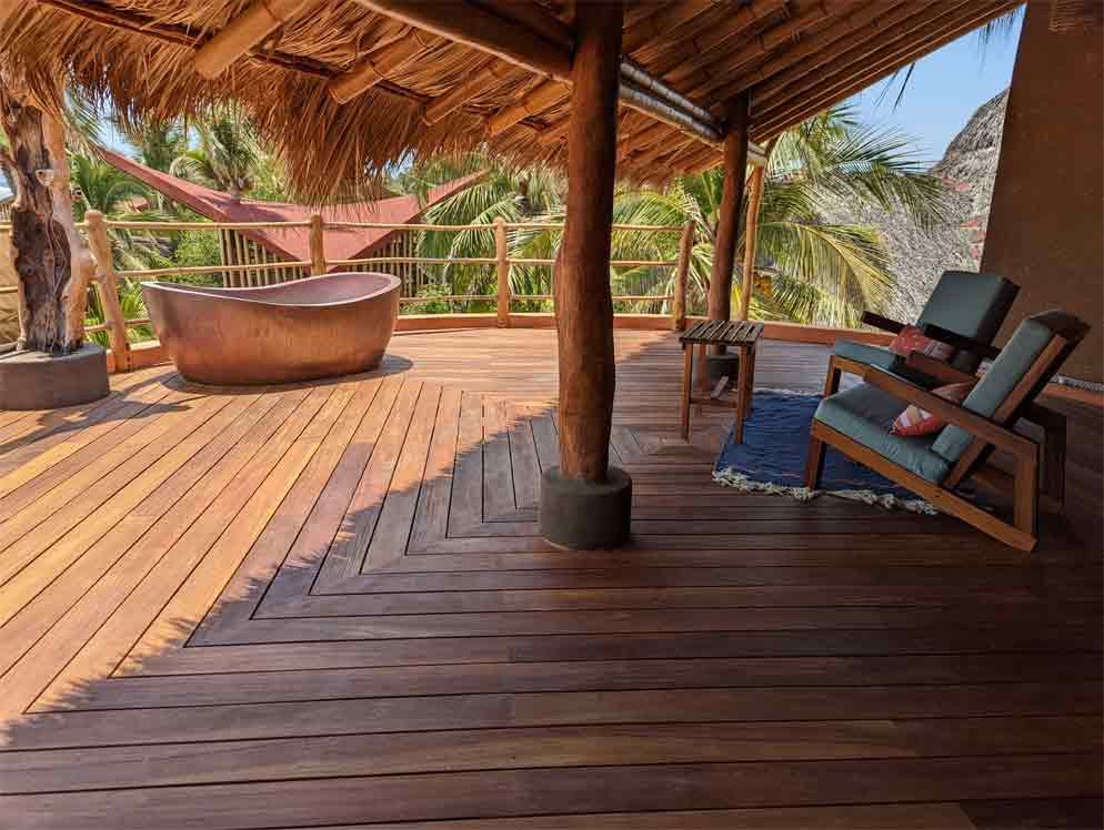 A wooden deck with a bathtub and chairs under a thatched roof.