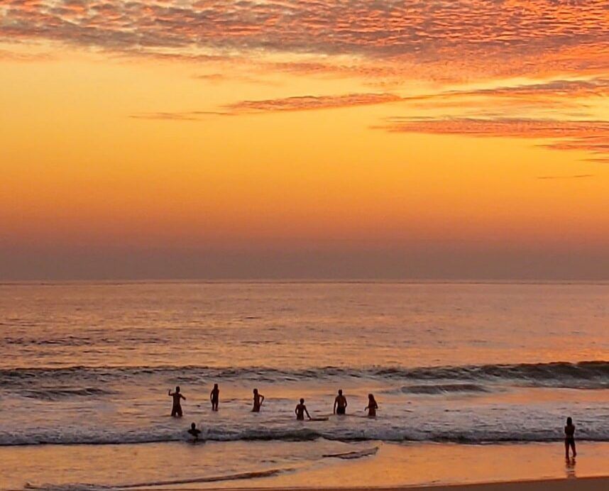 A group of people are standing in the ocean at sunset