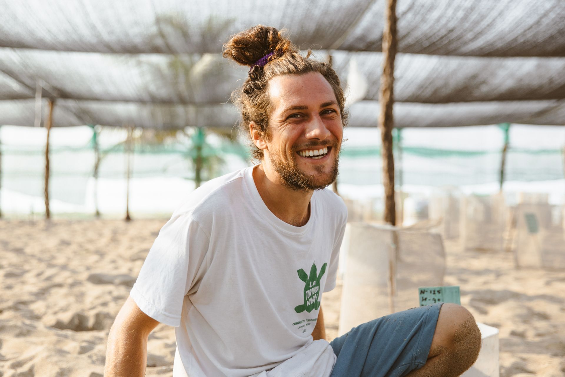 A man with a bun is sitting on the beach and smiling.