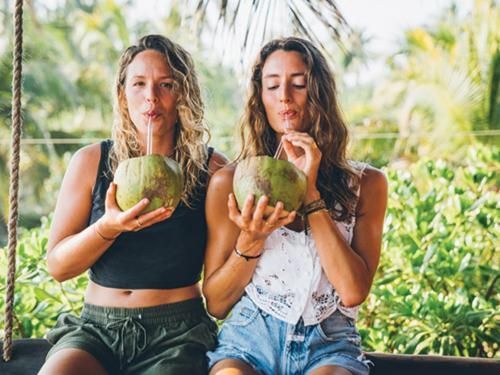 Two women are drinking coconut water through straws while sitting on a swing.