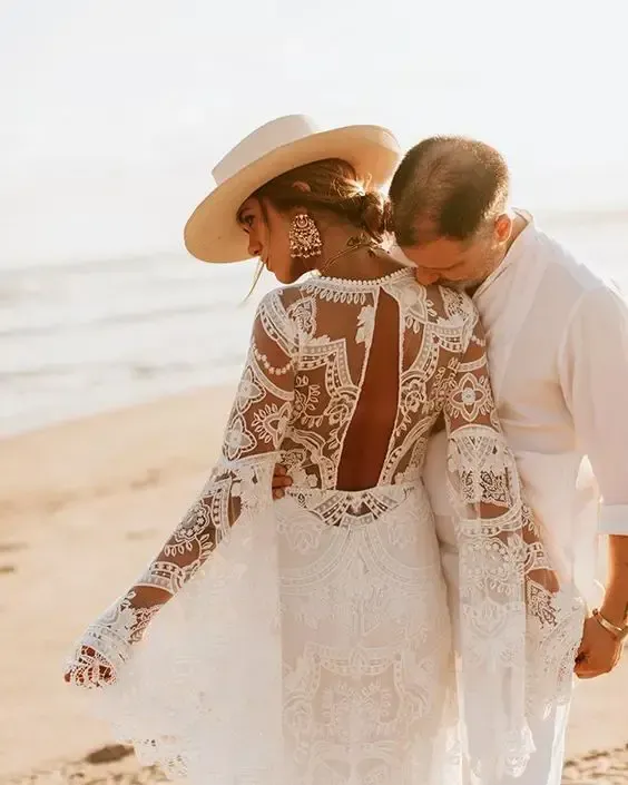 A bride and groom are walking on the beach . the bride is wearing a lace dress and a hat.
