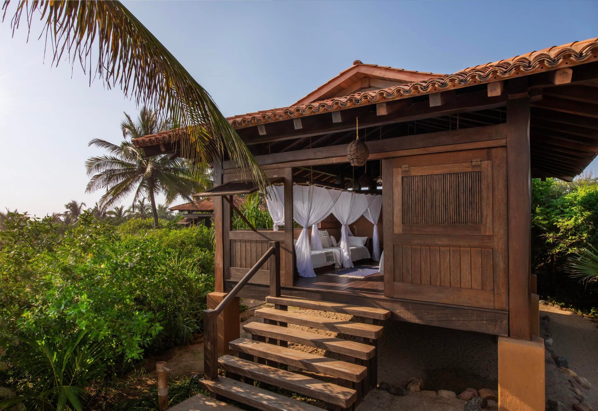 A small wooden house with stairs leading up to it is surrounded by palm trees.