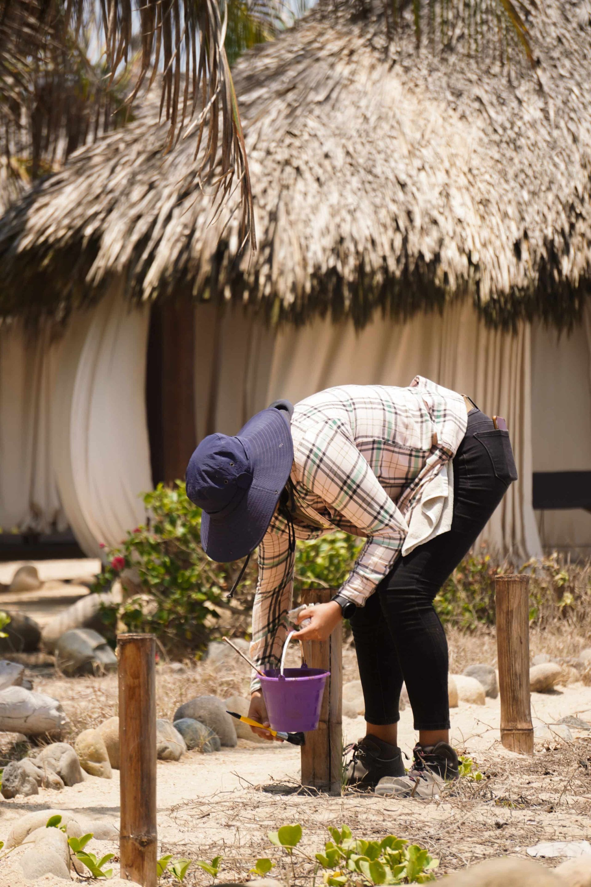 A woman is kneeling down in front of a thatched hut holding a bucket.