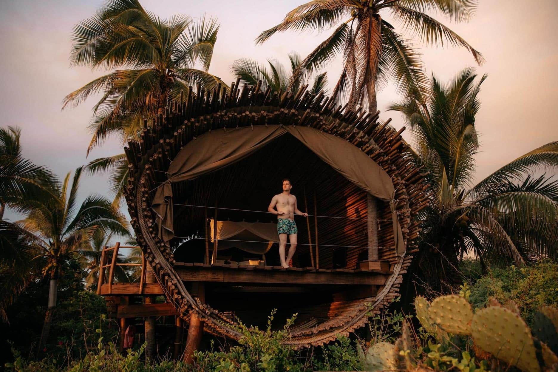 A man is standing on the balcony of a tree house surrounded by palm trees.