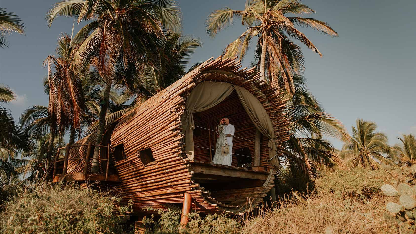 A woman in a white dress is standing in front of a wooden hut surrounded by palm trees.