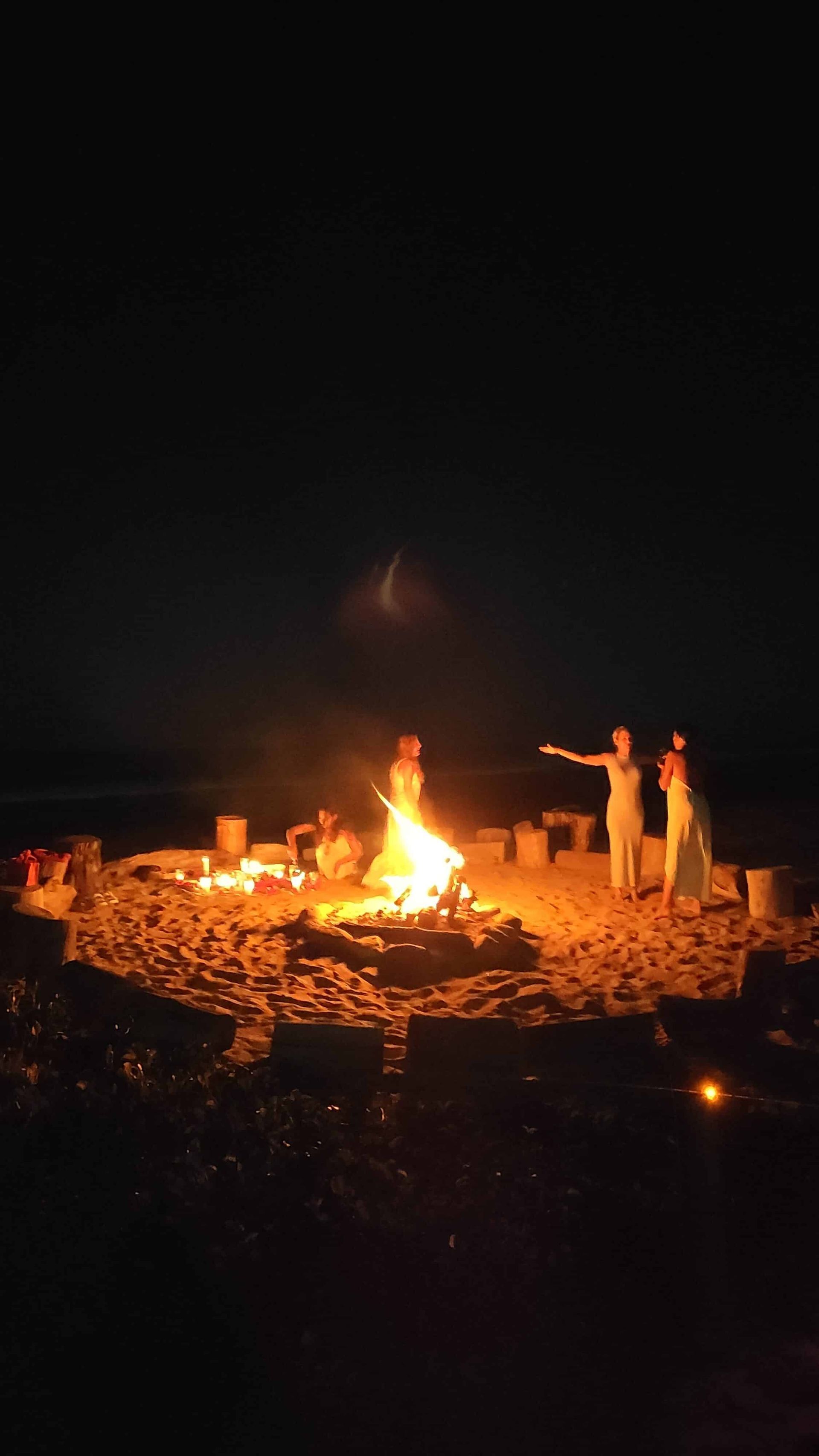 A group of people are standing around a fire on the beach at night.