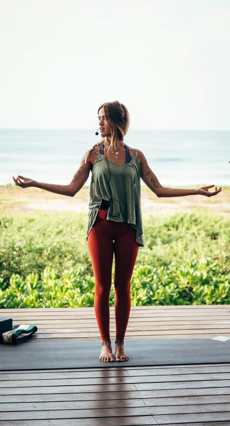A woman is standing on a yoga mat on a deck with her arms outstretched.
