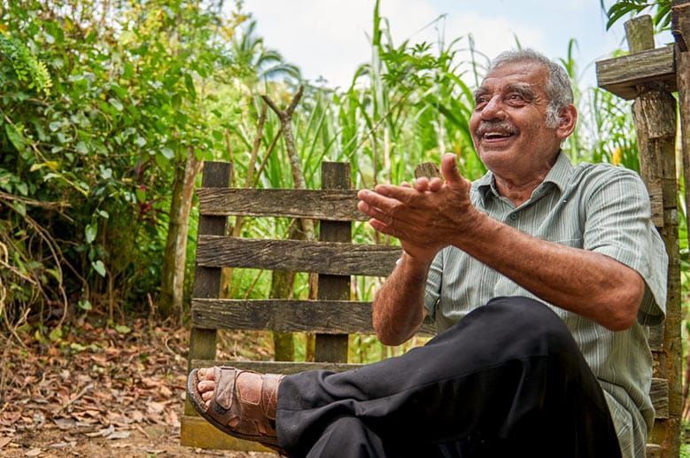 An elderly man is sitting on a wooden fence in a field.