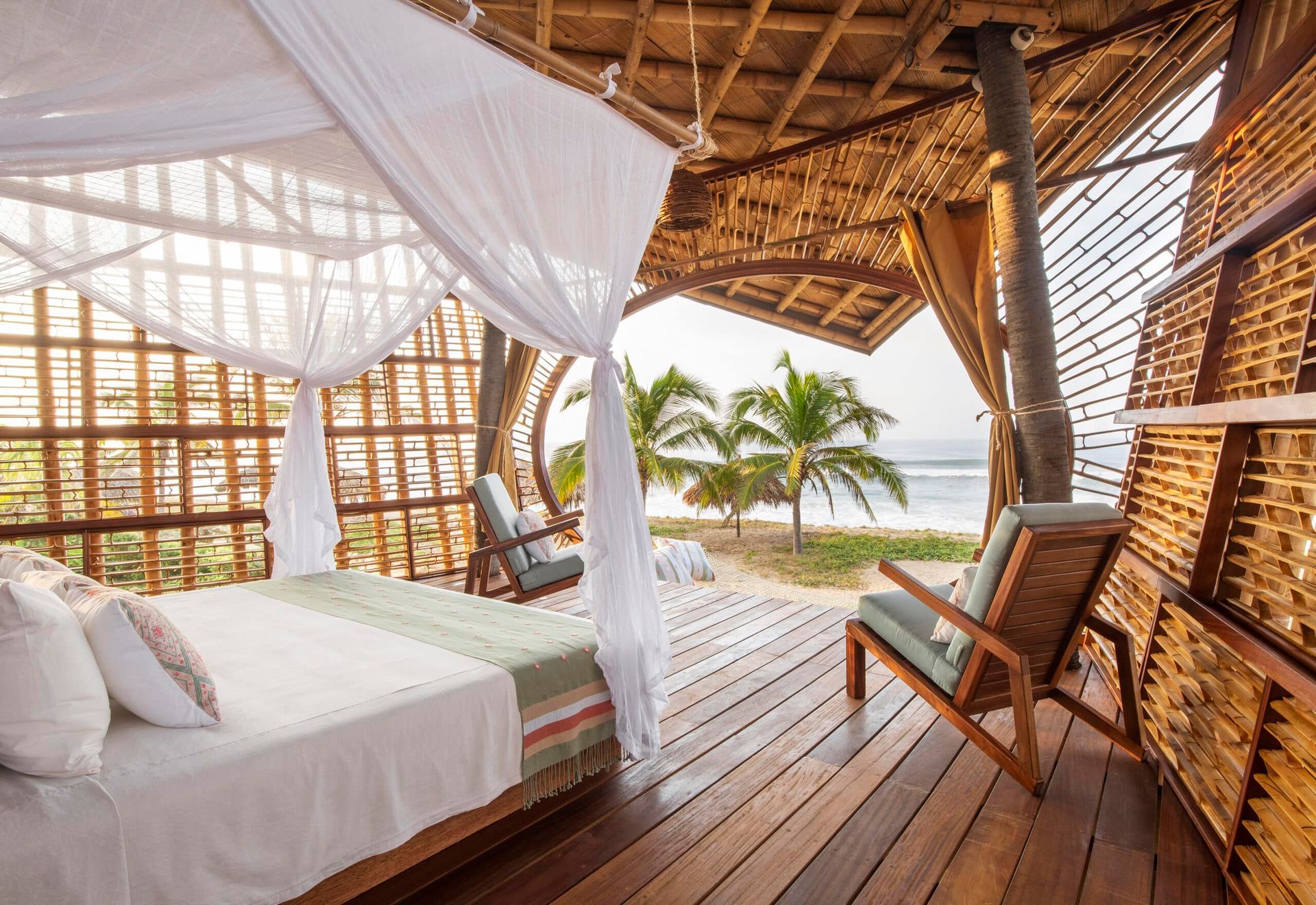 A bedroom with a canopy bed and chairs on a wooden deck overlooking the ocean.