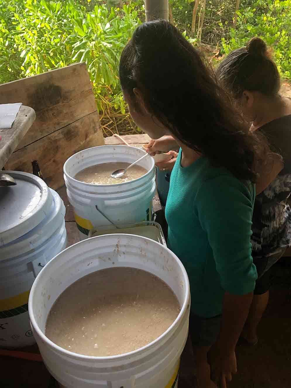 Two women are standing next to buckets of food on a table.