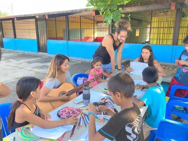 A group of children are sitting around a table with a woman playing a guitar.
