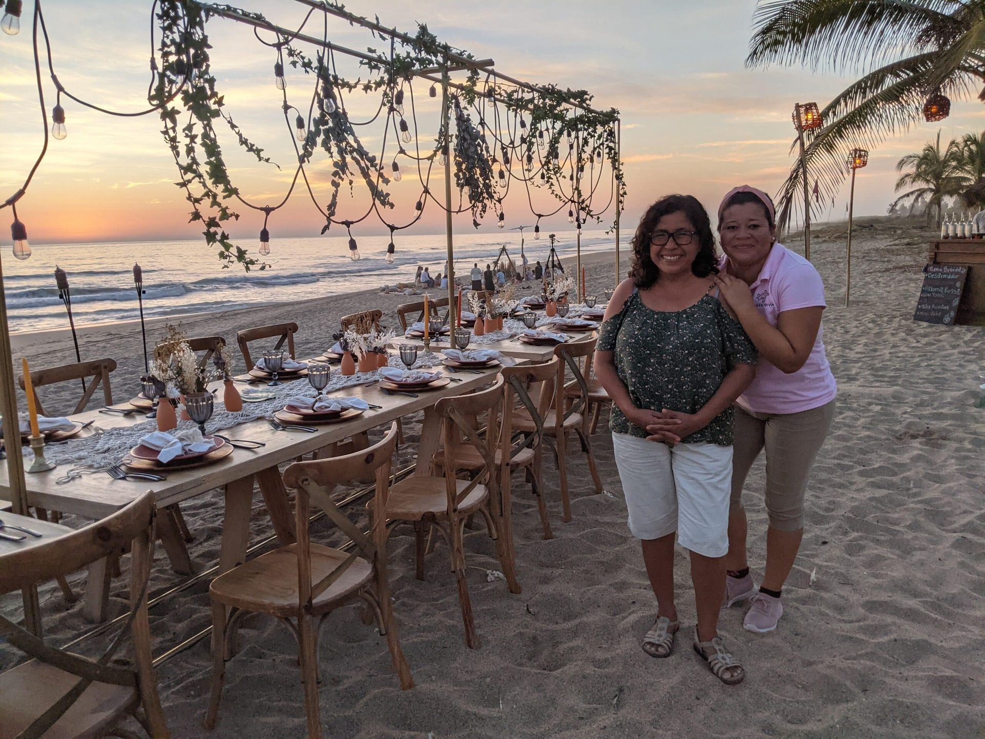 Two women are standing next to a long table on the beach.