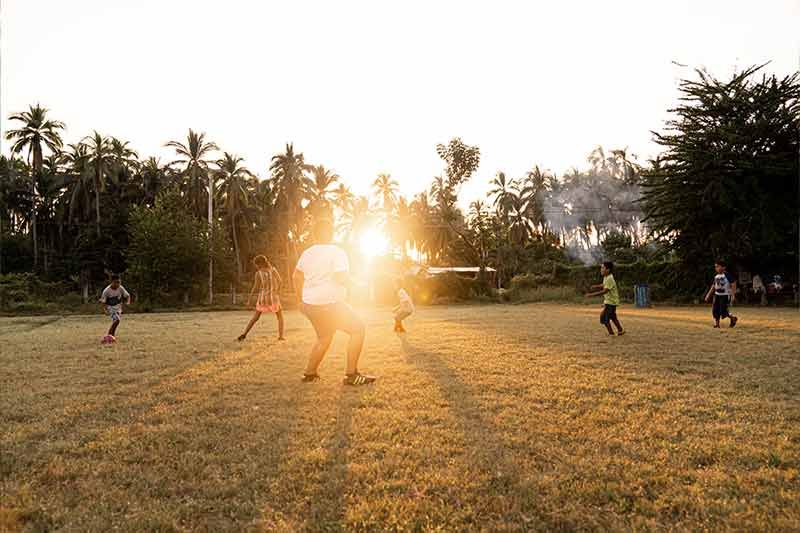 A group of people are playing soccer in a field with the sun shining through the trees.