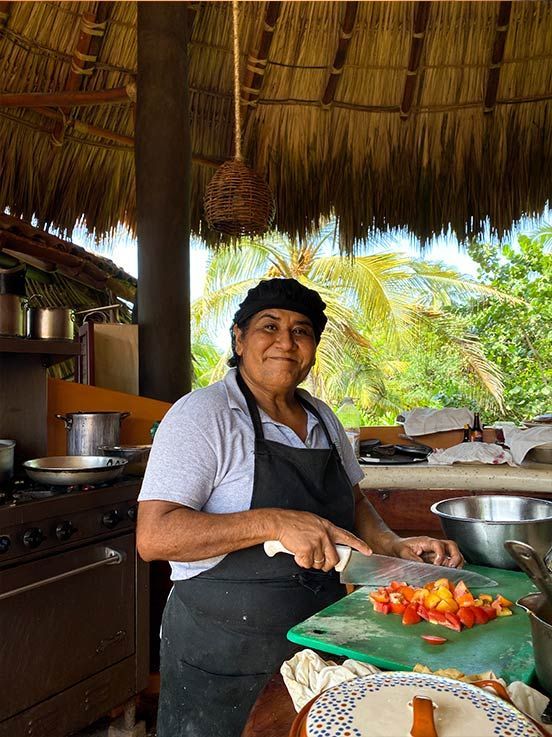 A woman is cutting vegetables on a cutting board in a kitchen under a thatched roof.