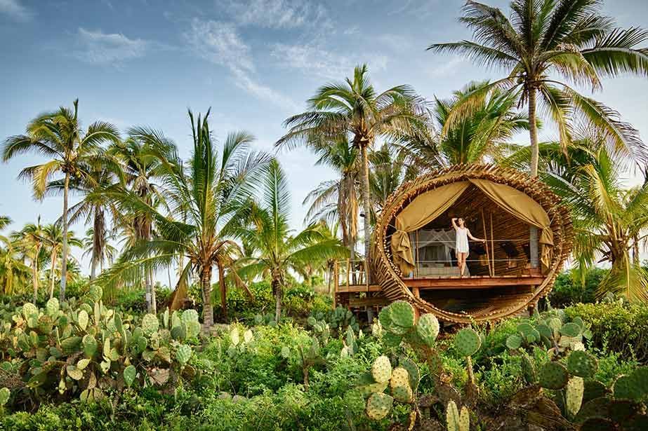A woman is standing in front of a tree house surrounded by palm trees.