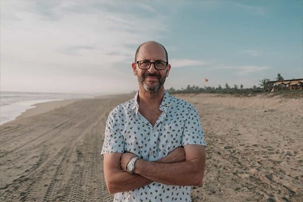 A man with glasses is standing on a beach with his arms crossed.