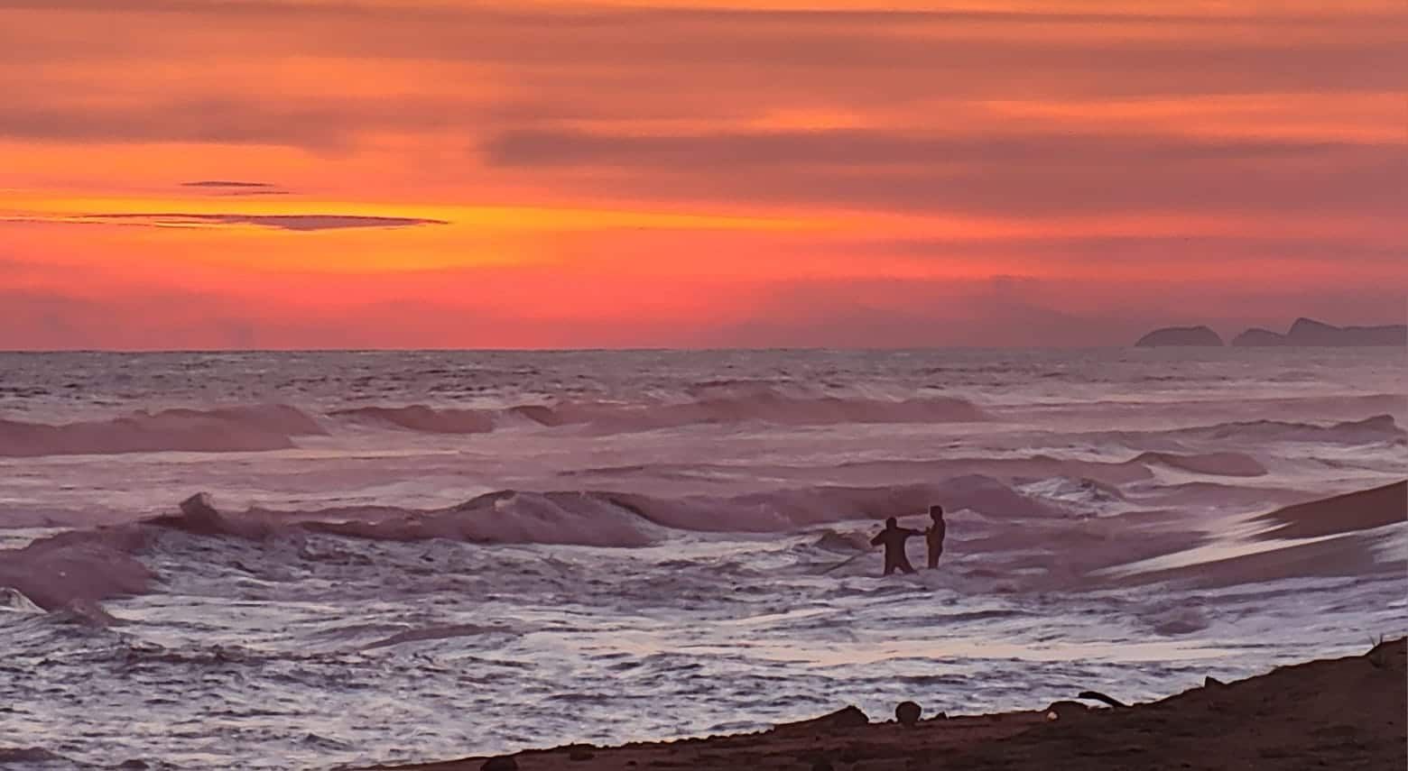A couple of people standing in the ocean at sunset.