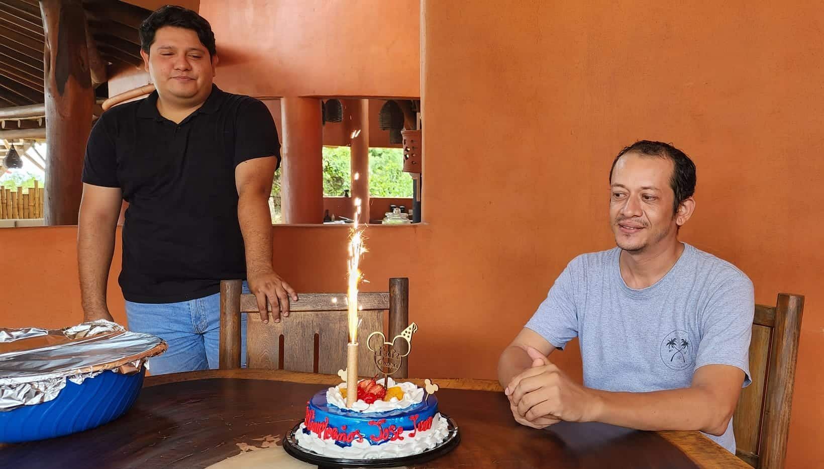 Two men are sitting at a table with a birthday cake and a candle.
