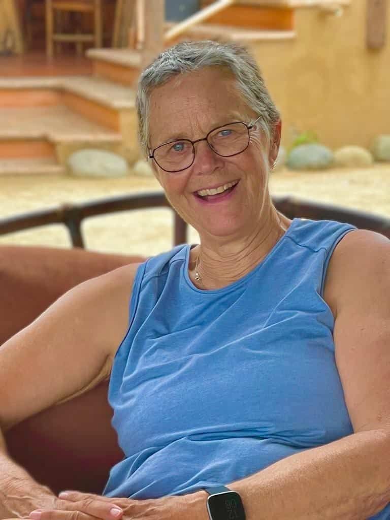 A woman wearing glasses and a blue tank top is sitting in a chair and smiling.