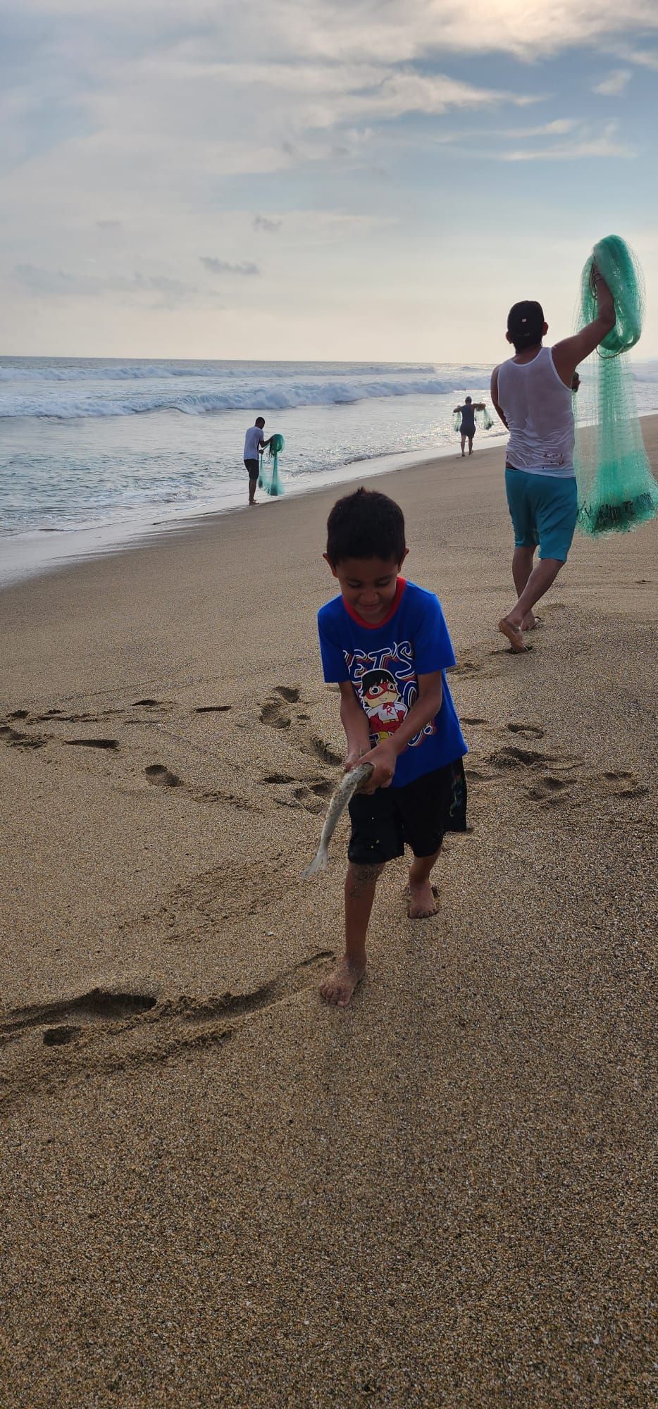 A little boy is running on the beach while a man holds a fishing net.