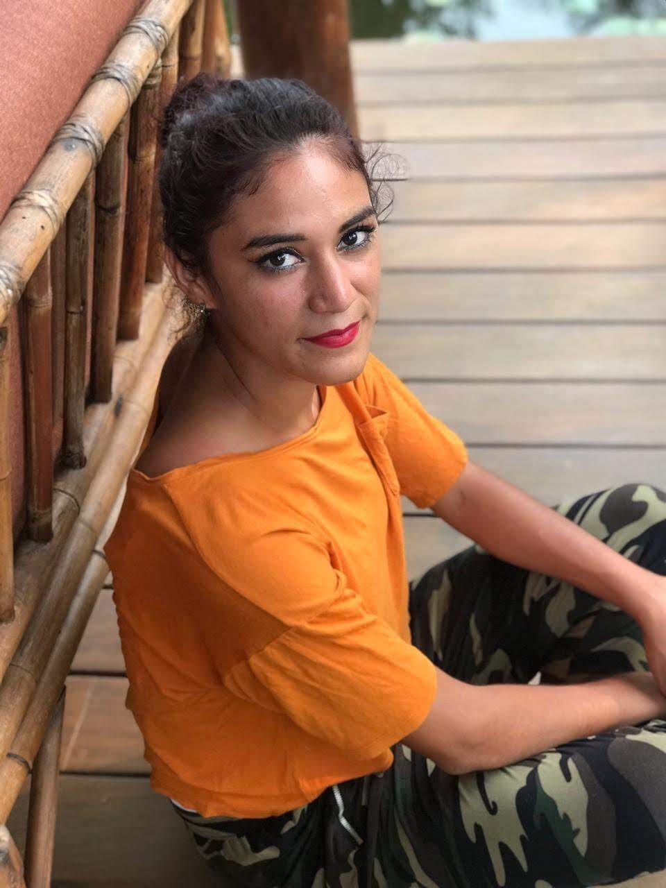 A woman in an orange top and camo pants is sitting on a wooden deck.