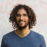 A man with curly hair and glasses is wearing a blue shirt and smiling.