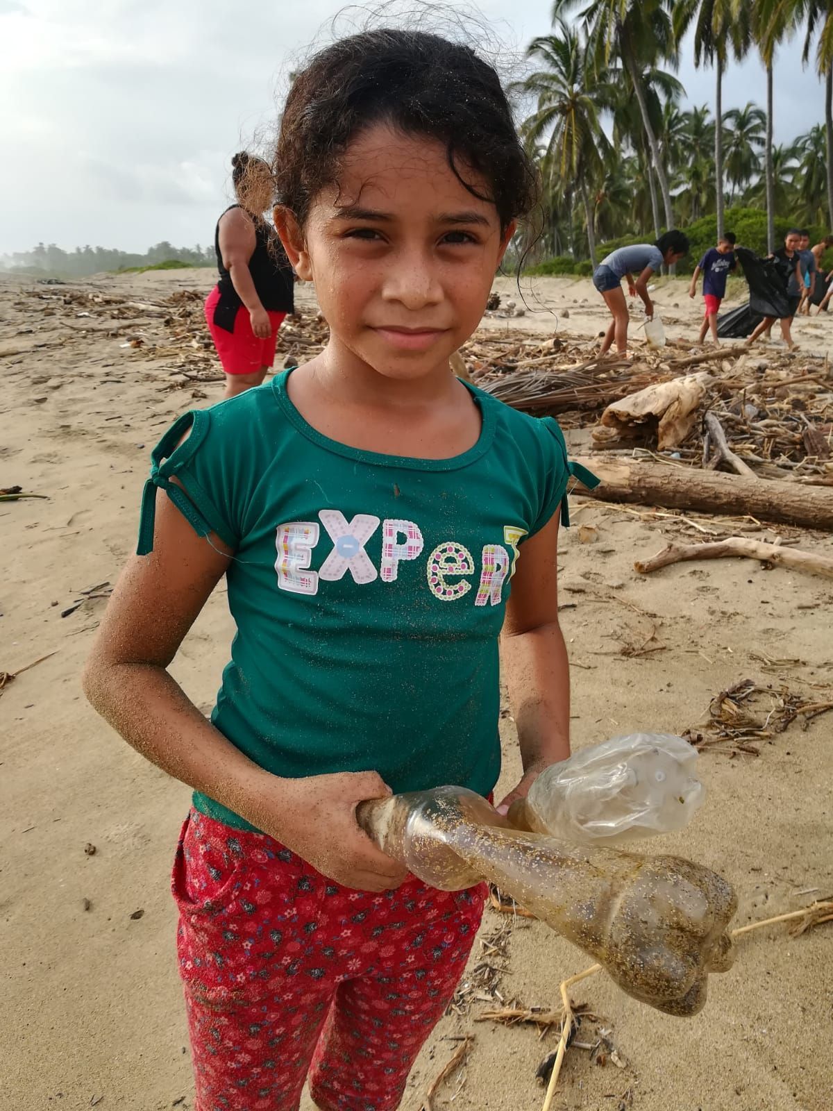A young girl in a green shirt is holding a plastic bottle on the beach.
