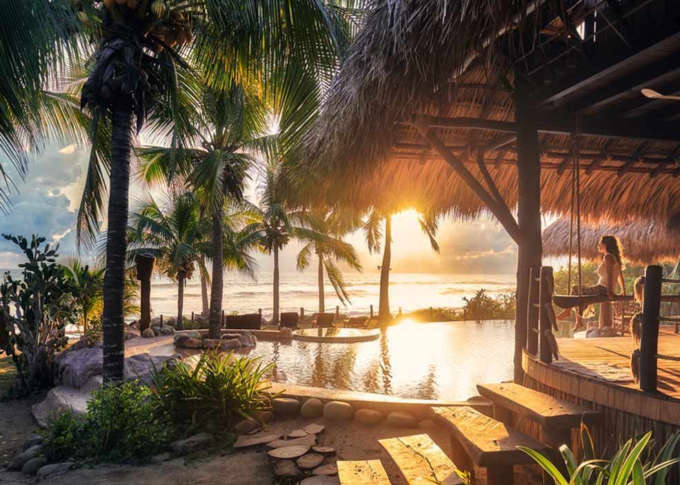 A couple of people are sitting under a thatched roof near a pool.