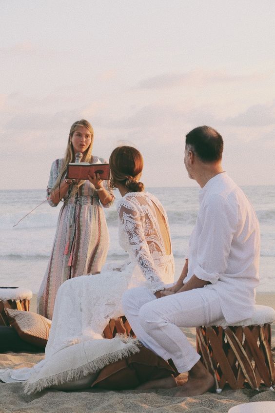 A bride and groom are sitting on the beach while a woman stands behind them.