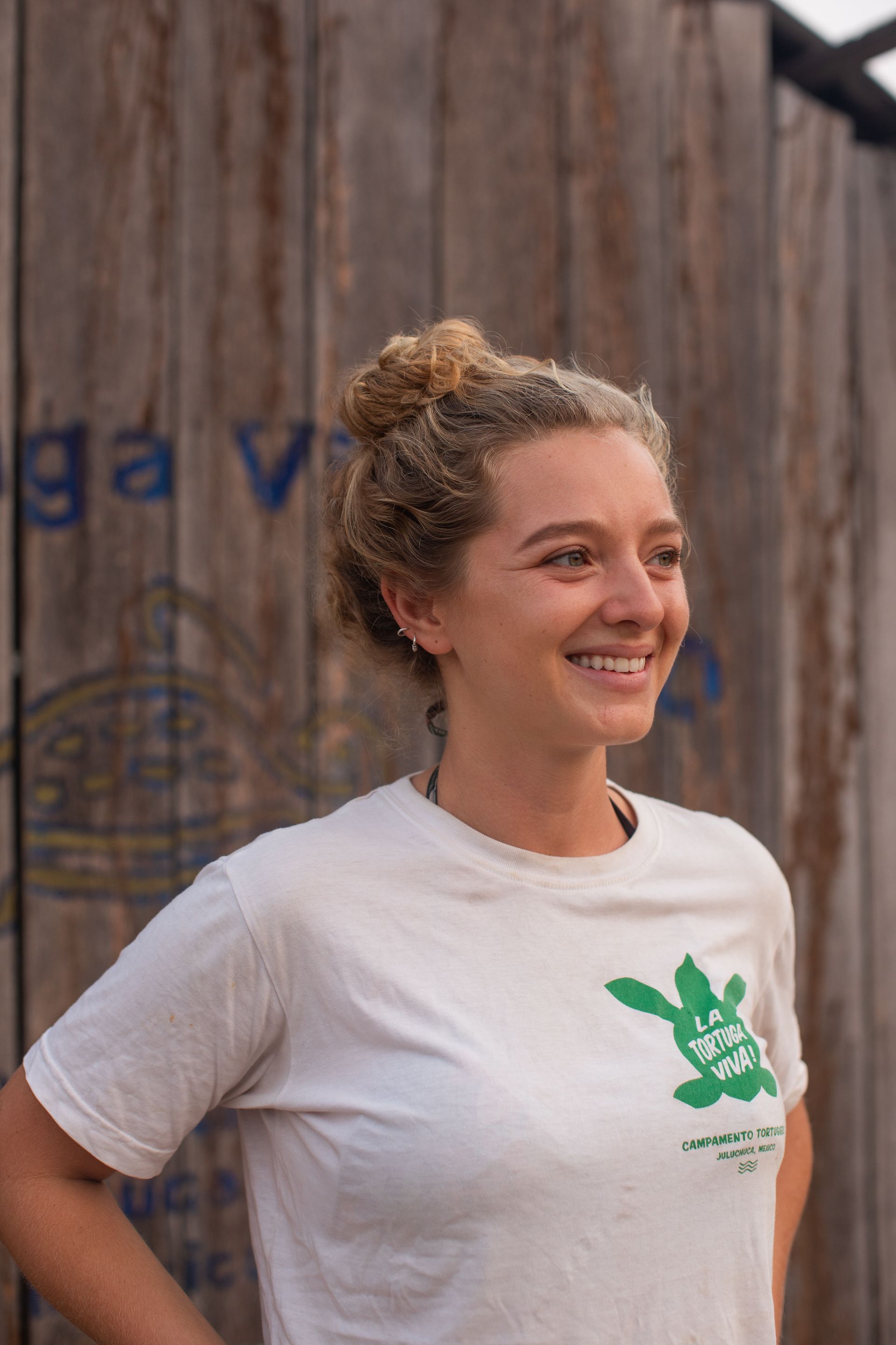 A woman in a white t-shirt is smiling in front of a wooden wall.