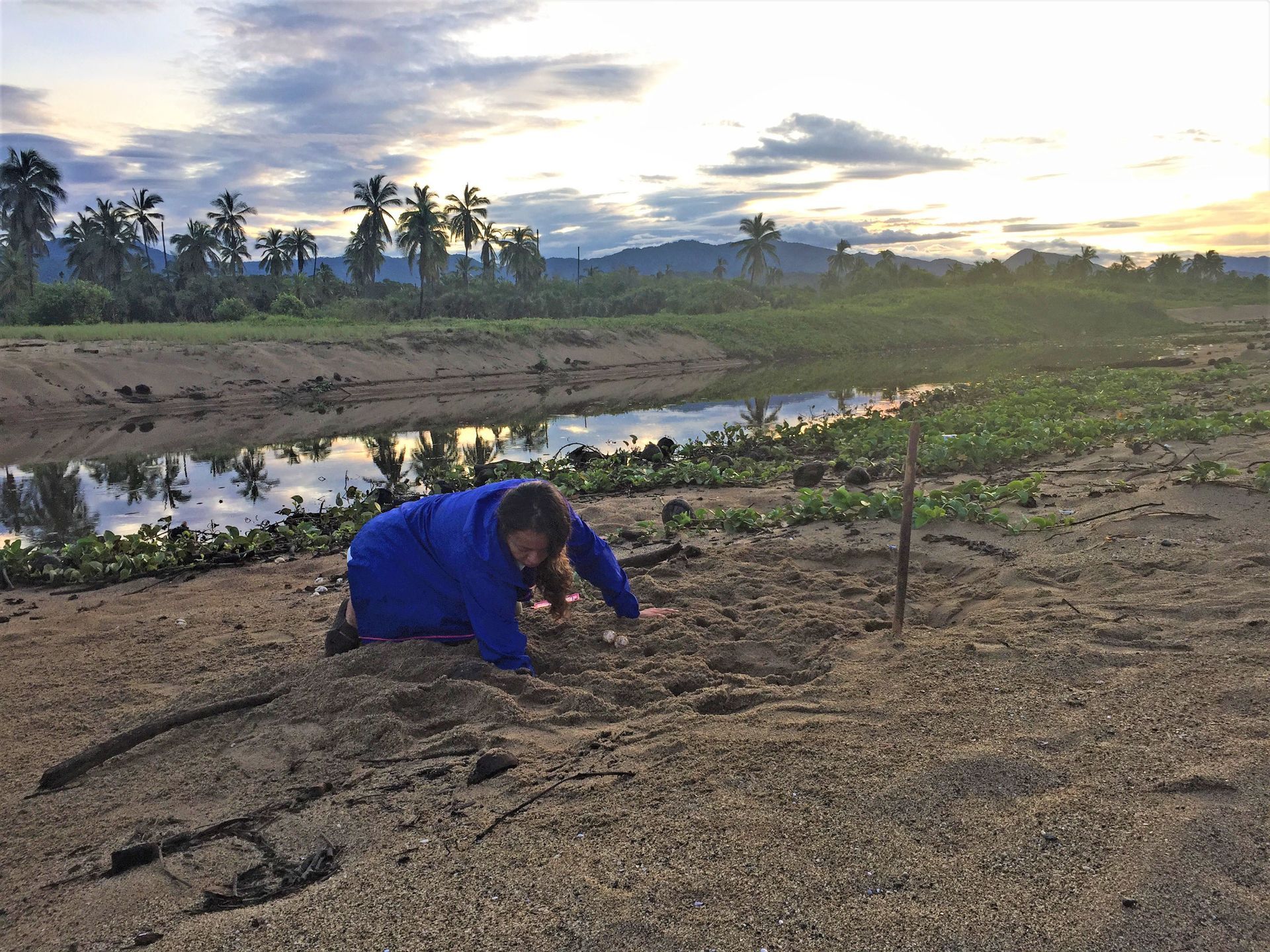 A person is kneeling in the dirt near a body of water.
