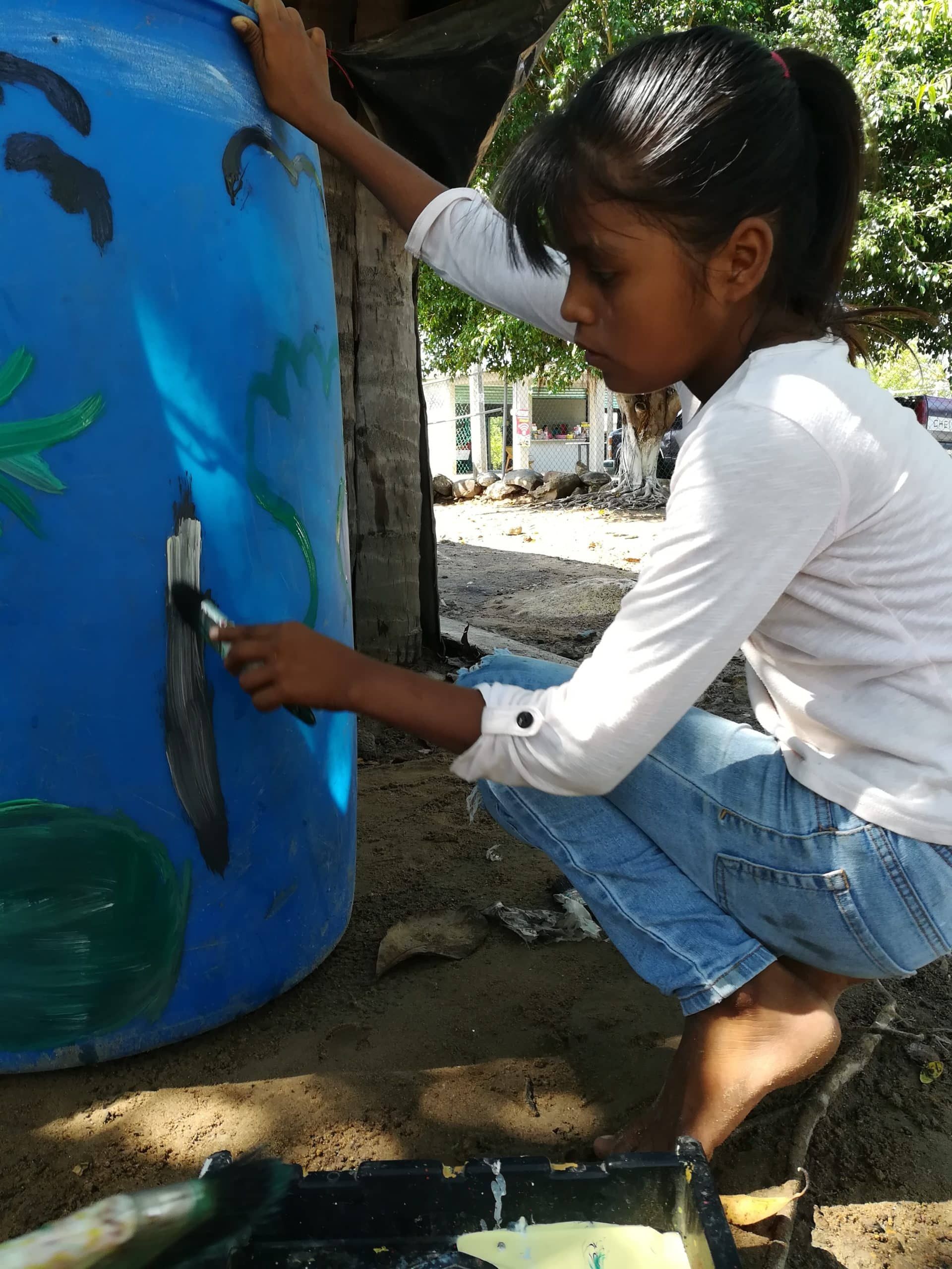 A young girl is painting a blue barrel with a brush