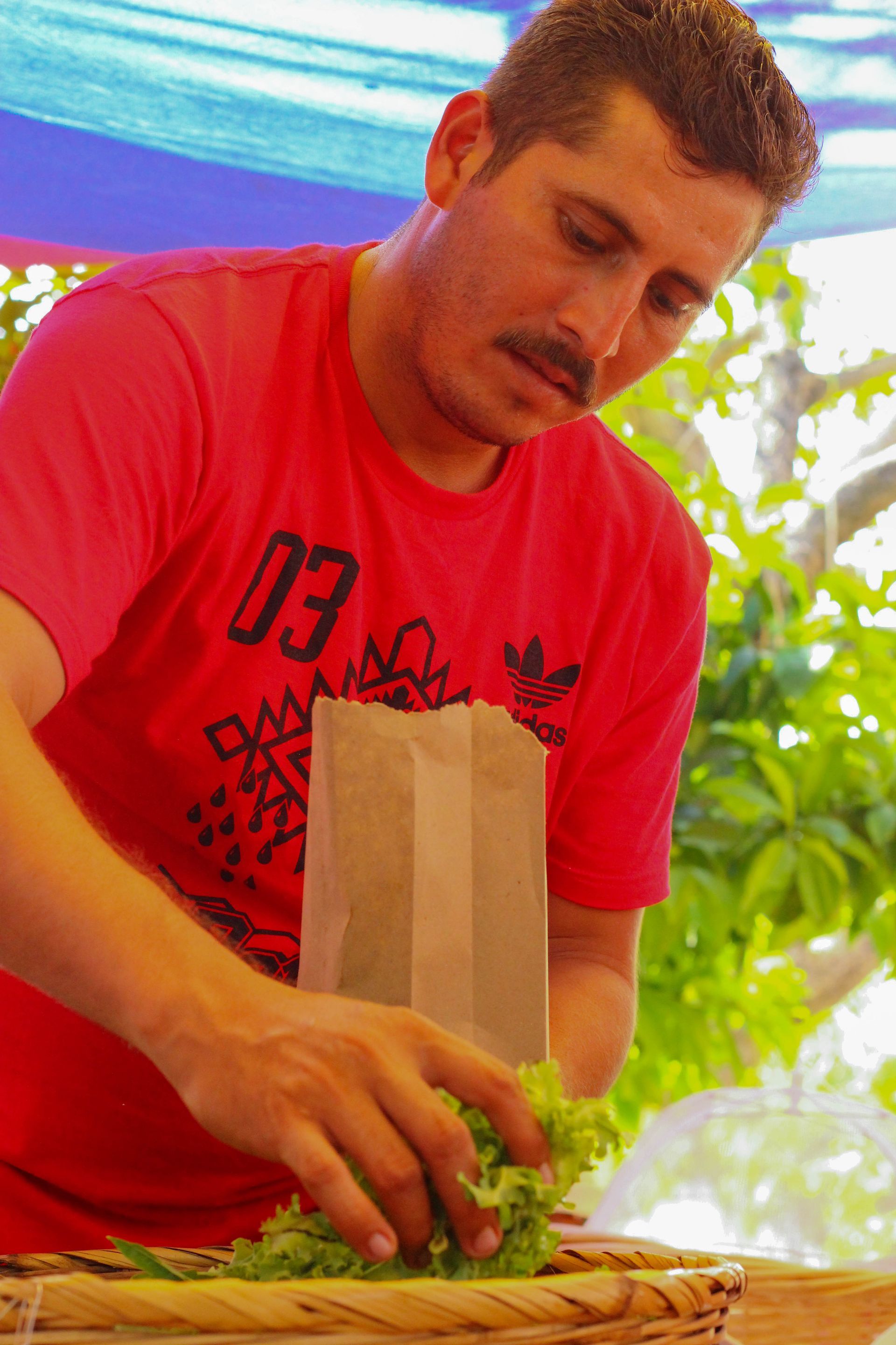 A man in a red shirt is putting lettuce on a sandwich.