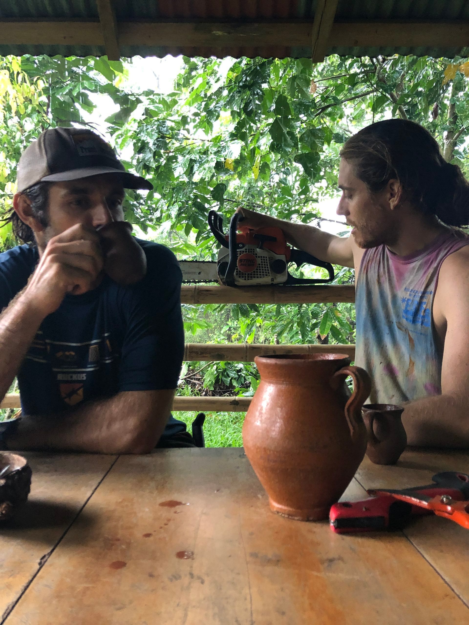 Two men are sitting at a table with a pot and a chainsaw.