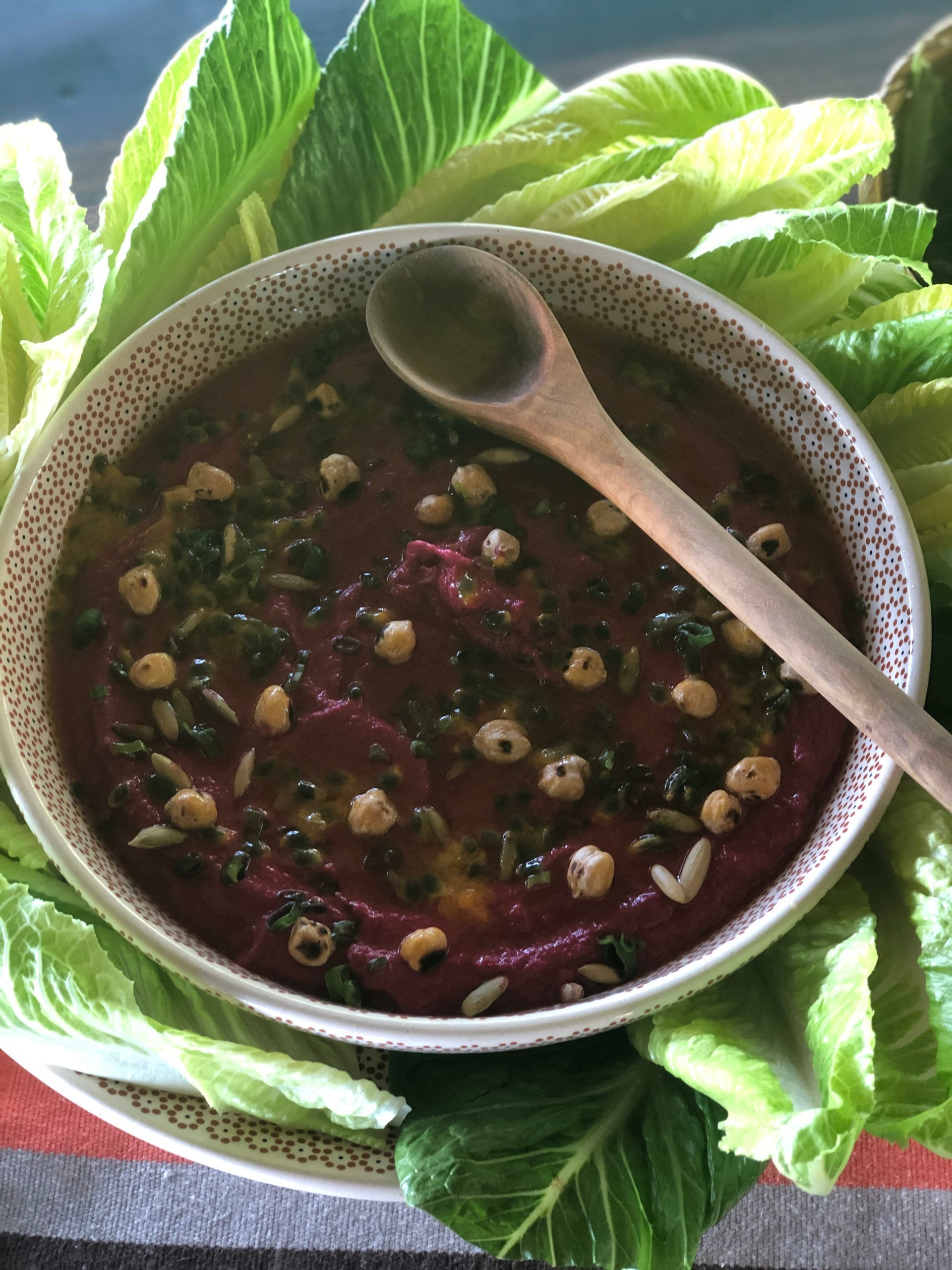 A bowl of soup with a wooden spoon in it is surrounded by lettuce leaves.