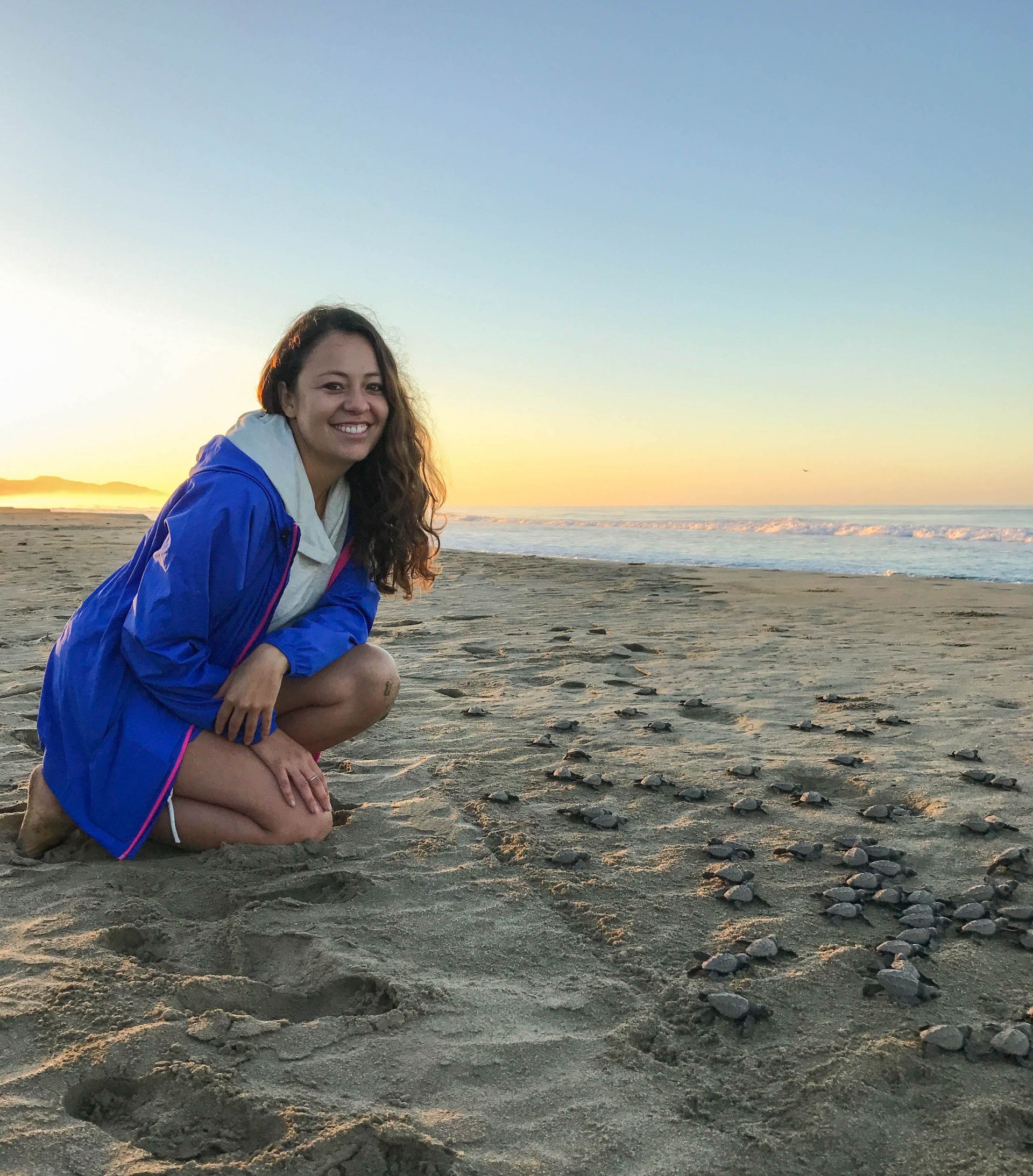 A woman in a blue coat is kneeling on the beach.